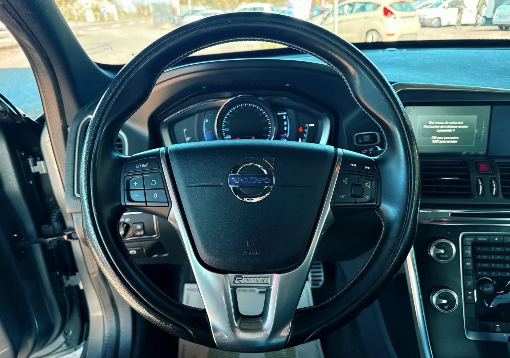 Vue rapprochée du volant noir du Volvo XC60 gris 2016 avec commandes intégrées et tableau de bord digital.