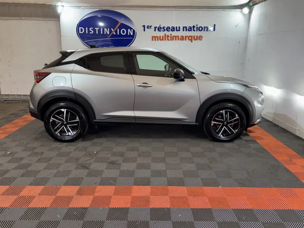Profil droit d’un Nissan Juke gris clair 2025, avec jantes noires et détails noirs contrastants, en intérieur showroom.