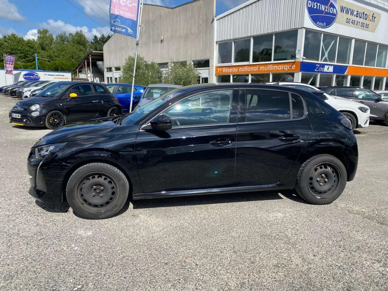 Peugeot 208 noire vue de profil côté gauche, stationnée sur parking devant un bâtiment commercial.