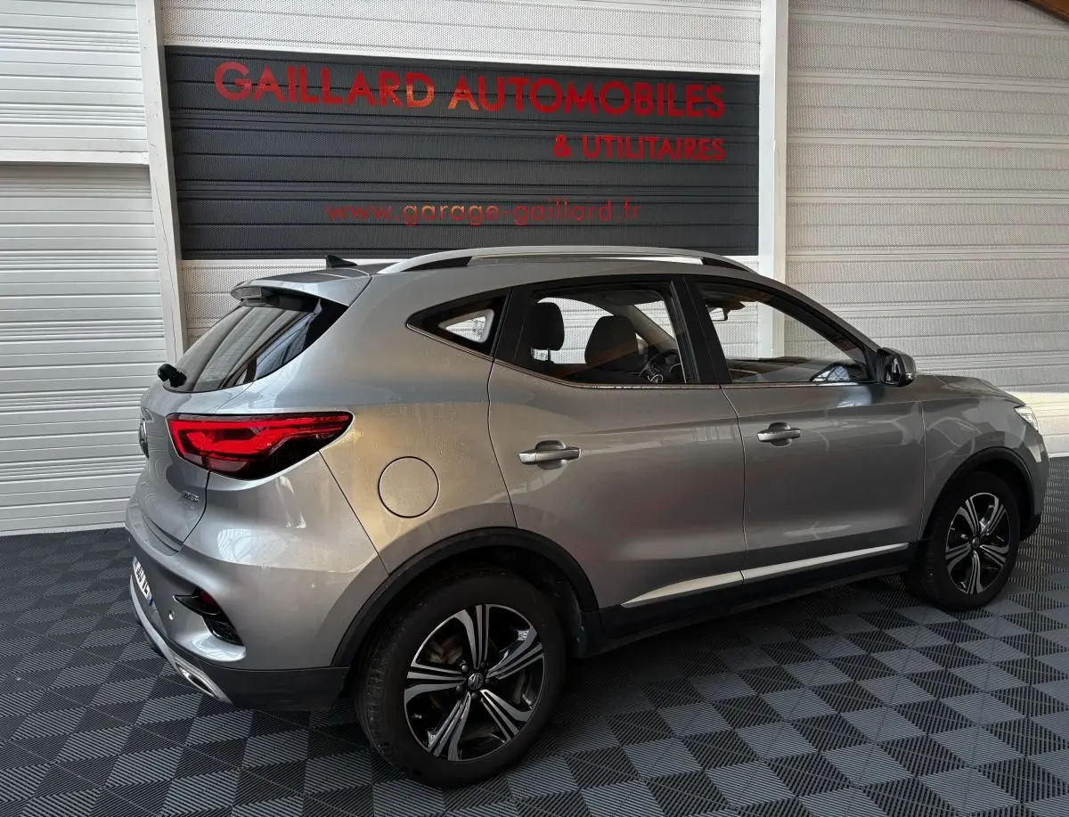 Vue 3/4 arrière côté droit d'un MG ZS gris clair 2022 avec jantes alliage et barres de toit chromées.