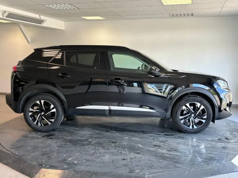 Profil côté gauche du Peugeot 2008 noir Perla Nera avec jantes alu et barres de toit visibles en intérieur showroom.