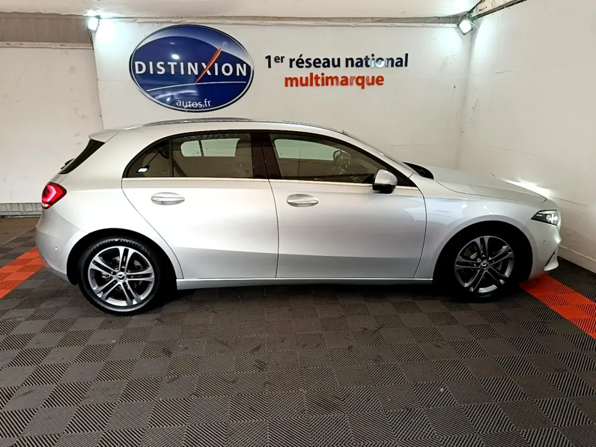 Vue de profil droit d'une Mercedes Classe A gris clair stationnée en intérieur sur sol à damiers noirs et rouges.