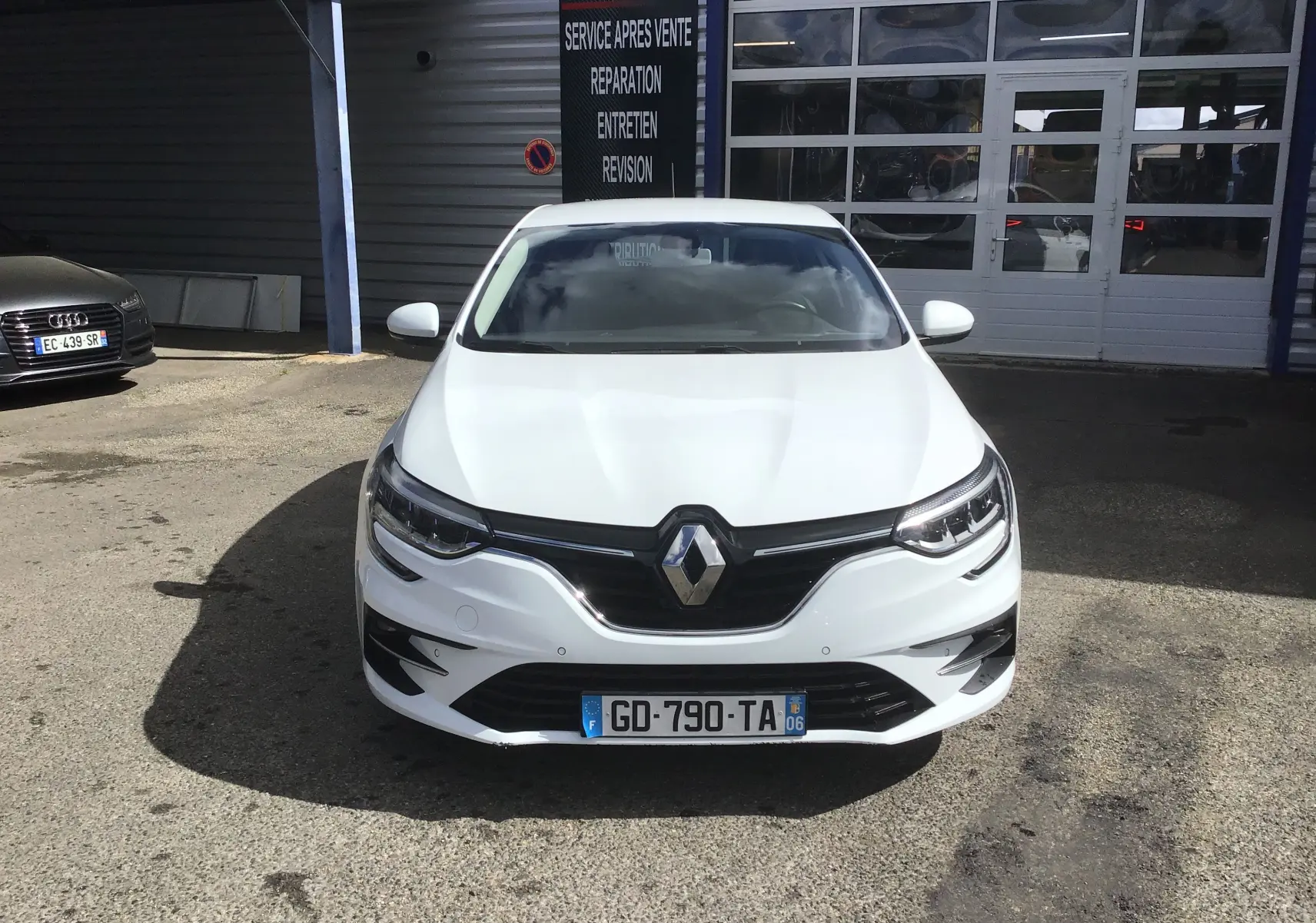Vue frontale d’une Renault Mégane IV Société blanche 2021, avec calandre noire et phares LED allumés, stationnée devant un garage.