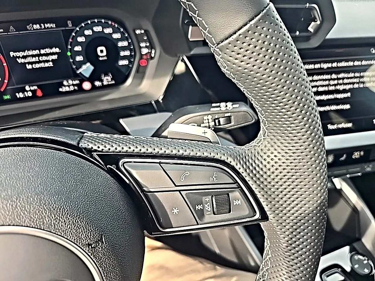 Gros plan sur le volant cuir perforé et les commandes multifonctions de l'Audi A3 Sportback gris Daytona, tableau de bord numérique visible.