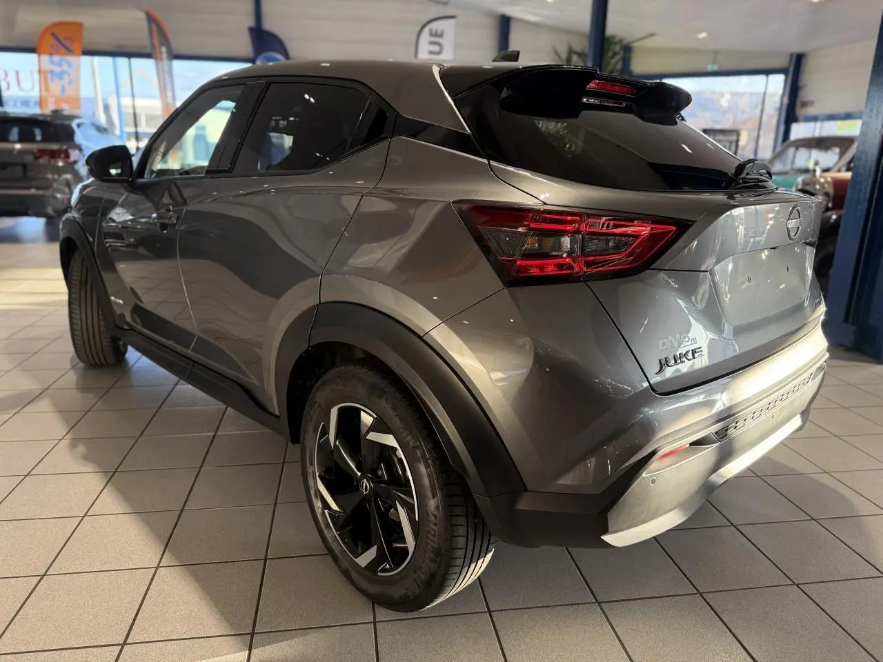 Vue 3/4 arrière droite du Nissan Juke Hybrid gris métallisé avec jantes alliage et toit noir en showroom.