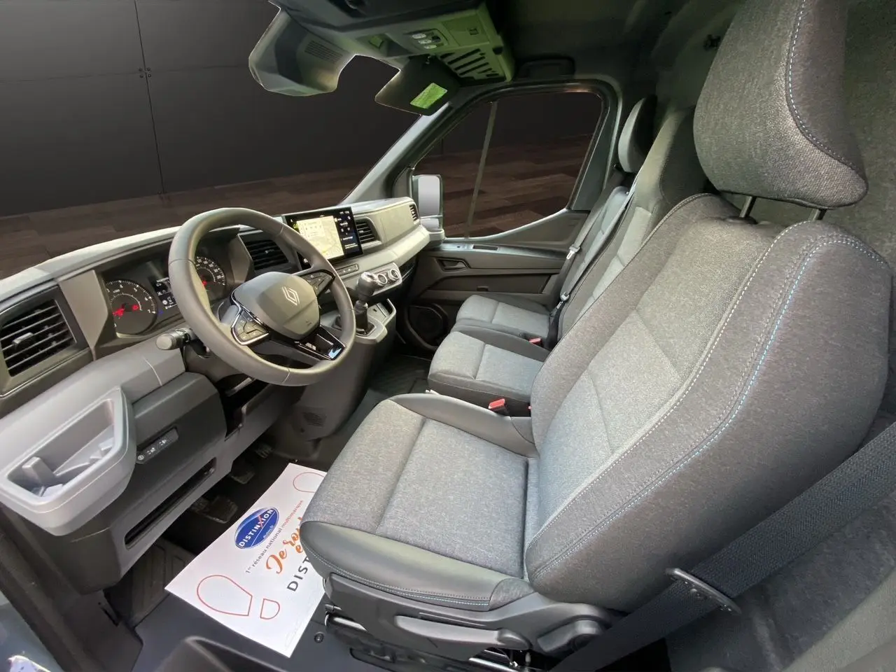 Vue intérieure côté conducteur du Renault Master 2025, montrant les sièges gris chiné avec surpiqûres bleues et le tableau de bord moderne.