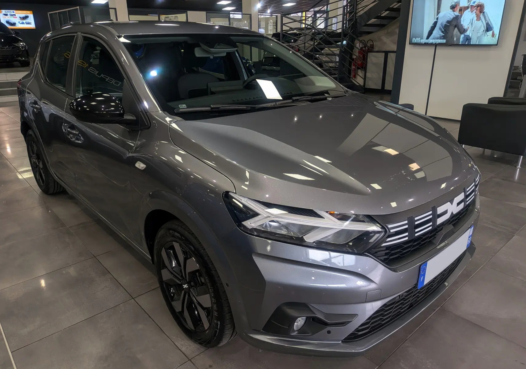Vue 3/4 avant d'une Dacia Sandero TCe 90 CH Journey gris schiste avec jantes noires et calandre lumineuse en showroom.