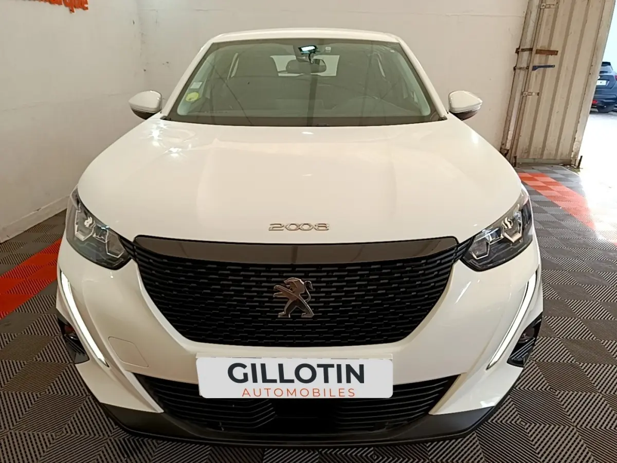 Vue avant d'une Peugeot 2008 blanche avec calandre noire et phares LED, stationnée en intérieur sur sol damier.