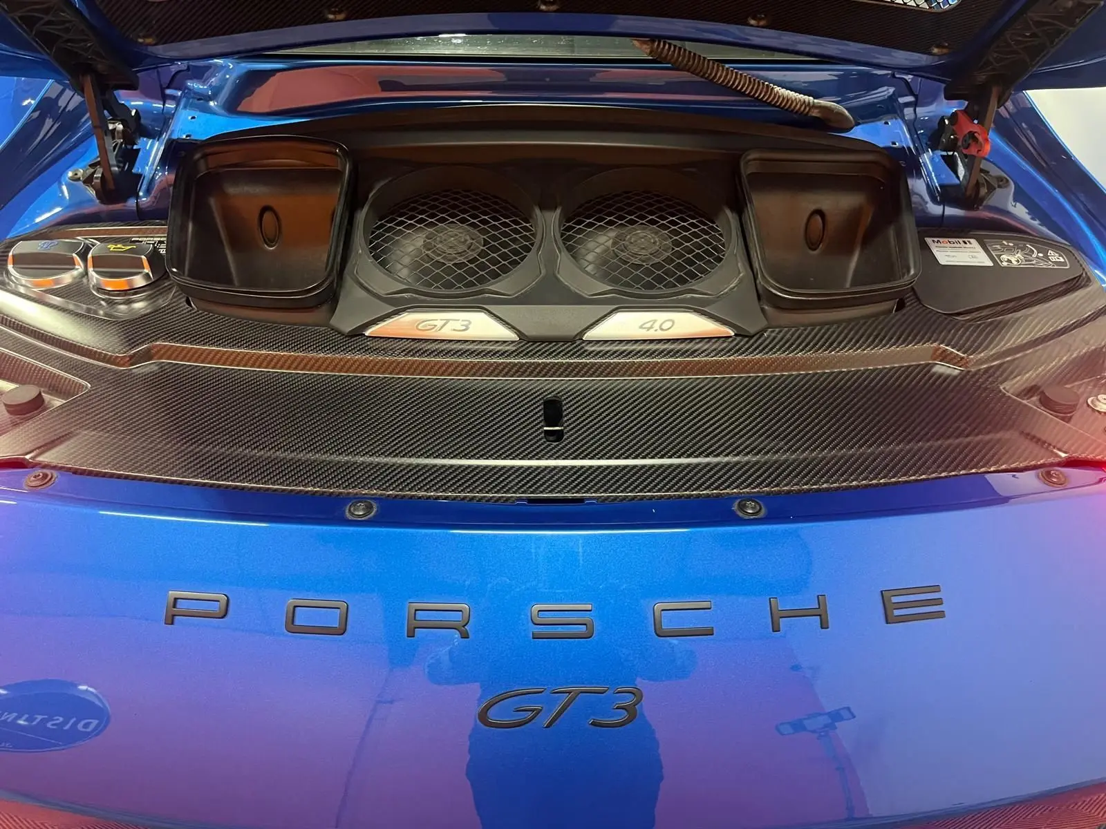 Vue rapprochée du compartiment moteur arrière d’une Porsche 911 GT3 bleu saphir avec détails en carbone et logos GT3 4.0 visibles.
