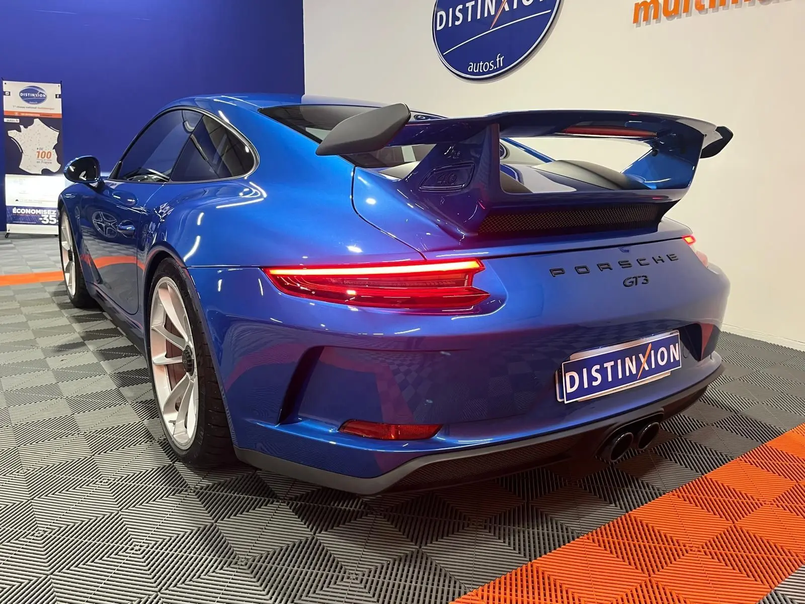 Vue 3/4 arrière droite d'une Porsche 911 GT3 bleu saphir avec aileron arrière et feux LED allumés en intérieur showroom.