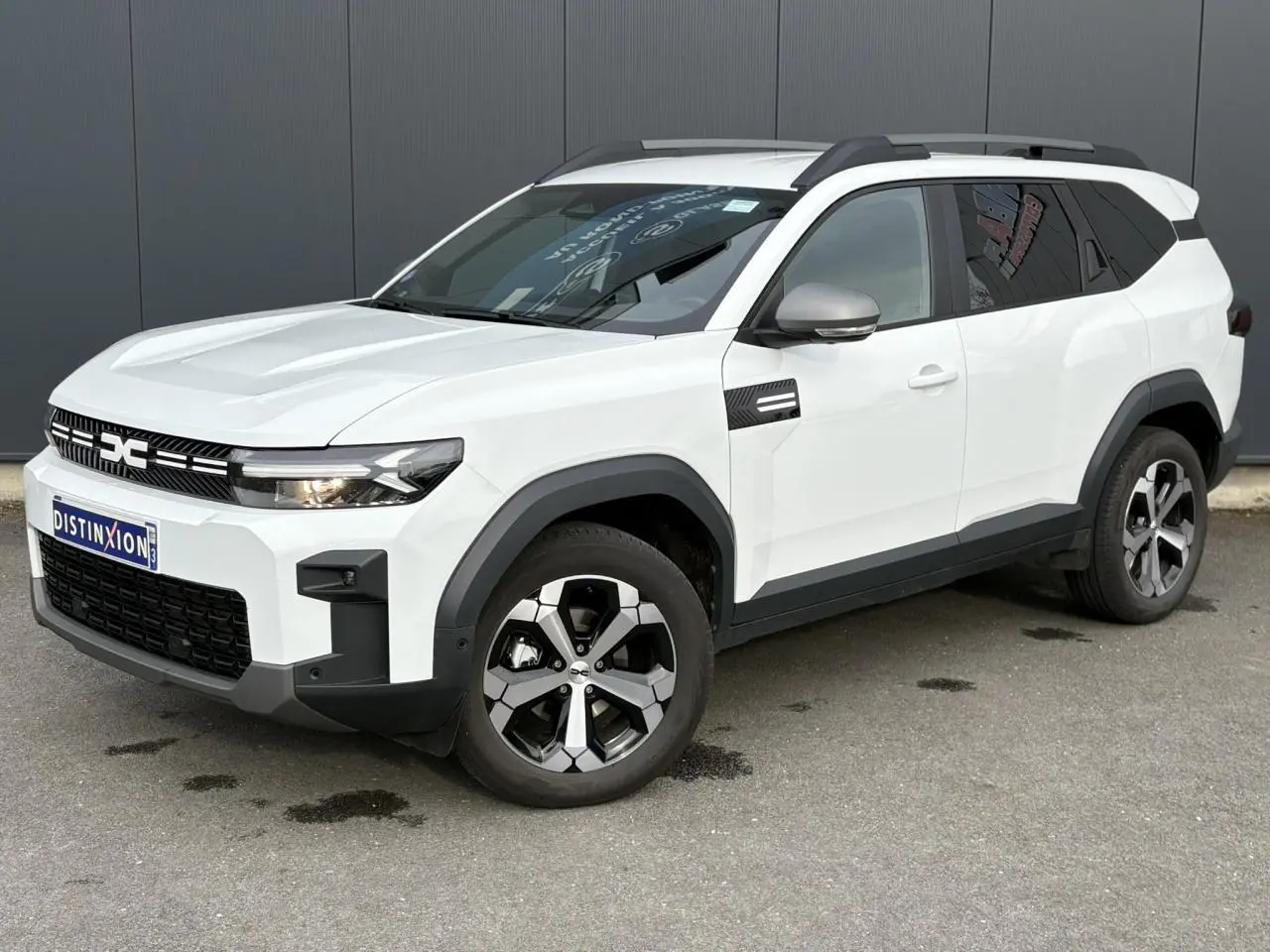 Dacia Bigster blanc Banquise en 3/4 avant droit, avec barres de toit gris et jantes alliage 18 pouces diamantées.