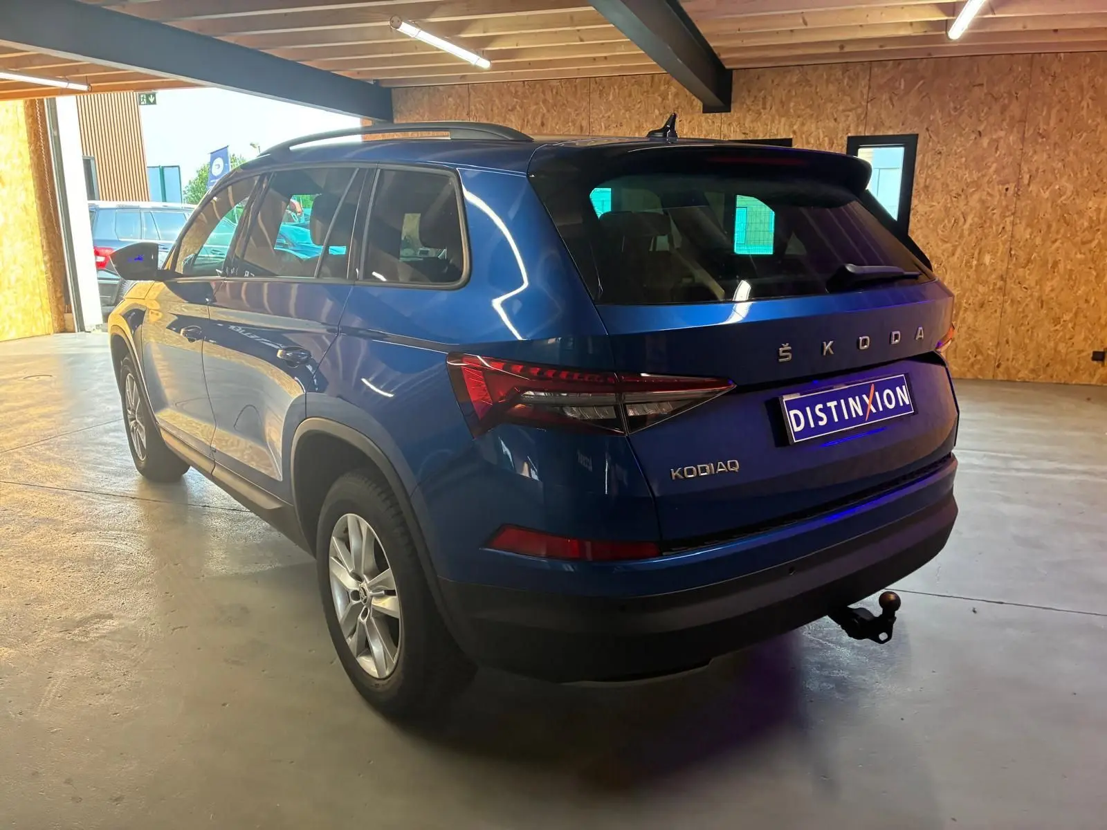 Vue 3/4 arrière droite d'un Skoda Kodiaq bleu 2023 avec attelage de remorque, dans un garage lumineux.
