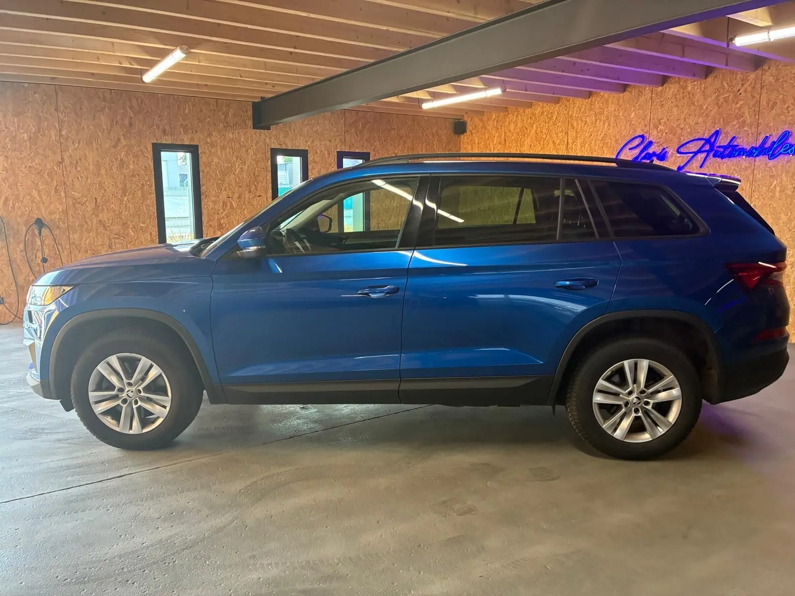 Vue de profil côté gauche d'un Skoda Kodiaq bleu 2023 stationné en intérieur avec jantes alliage et vitres teintées.