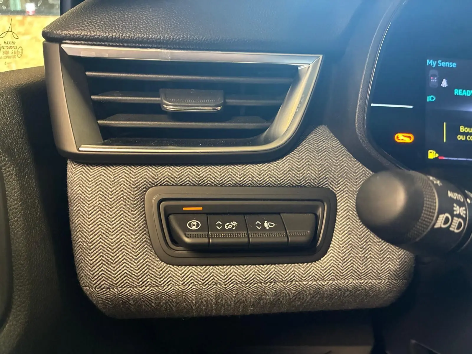 Détail intérieur côté gauche de la planche de bord de la Renault Clio 2025, tissu gris à chevrons avec commandes d’éclairage.