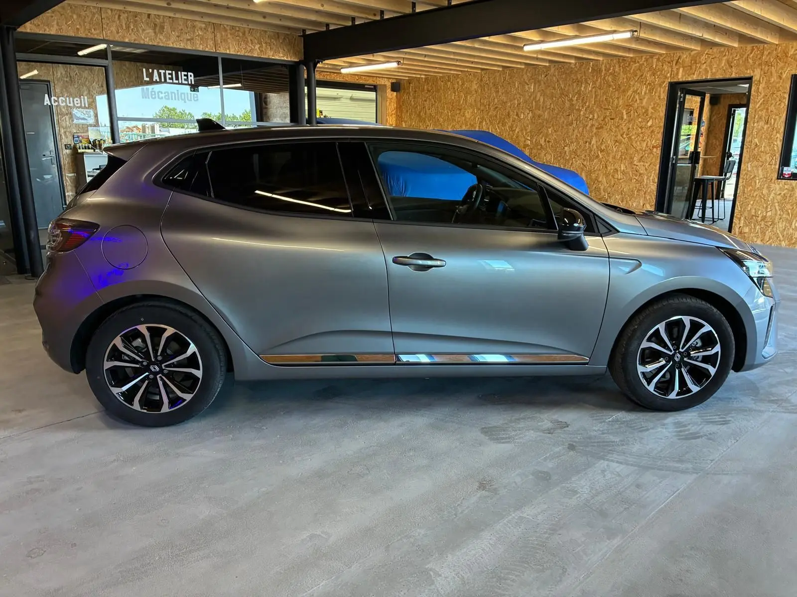Profil côté gauche de la Renault Clio 2025 gris schiste, avec jantes noires et détails chromés, en intérieur d'atelier.