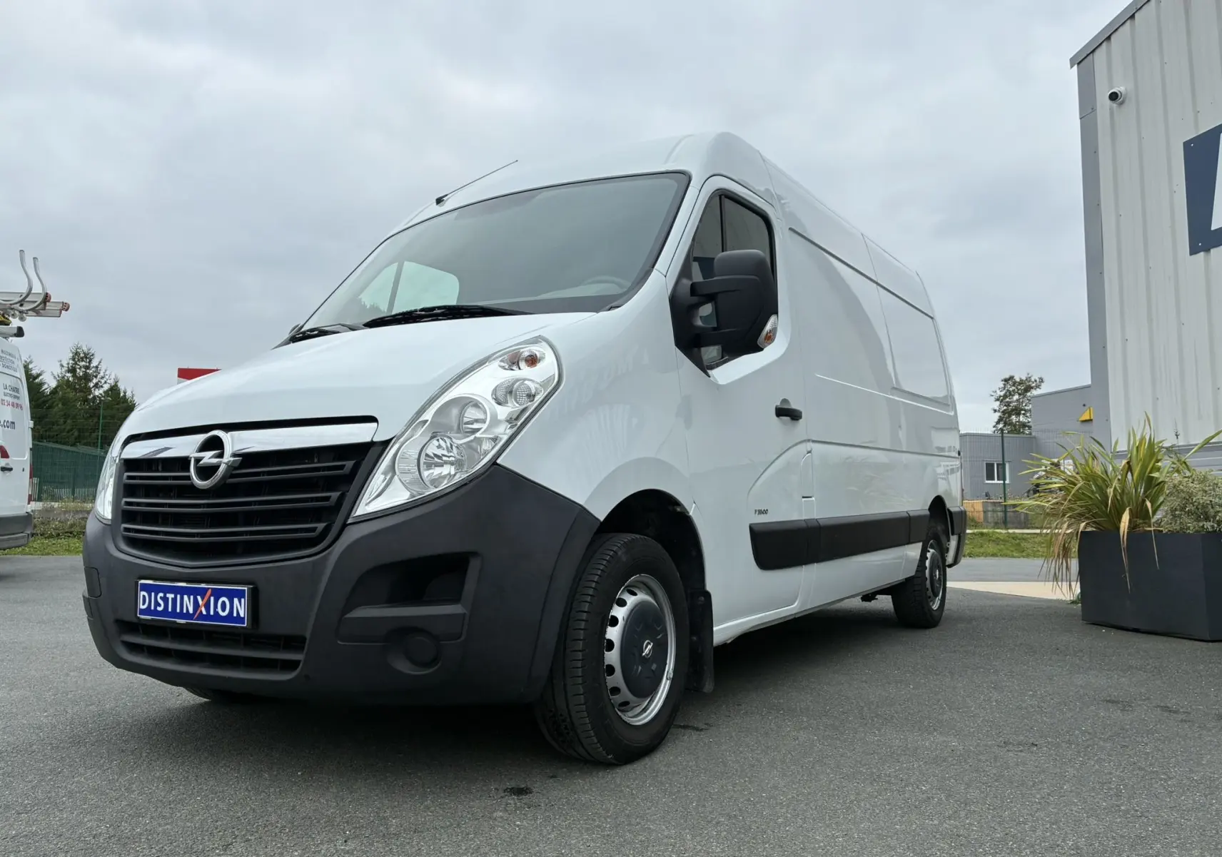Fourgon Opel Movano blanc vu en 3/4 avant droit, avec pare-chocs noir et jantes acier, stationné sur un parking.