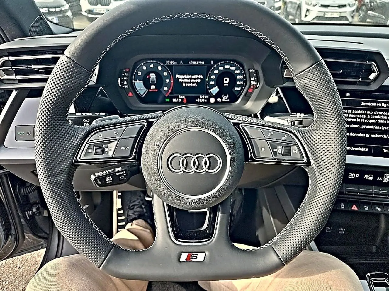 Vue rapprochée du volant cuir perforé noir avec logo Audi et badge S, tableau de bord digital en arrière-plan.