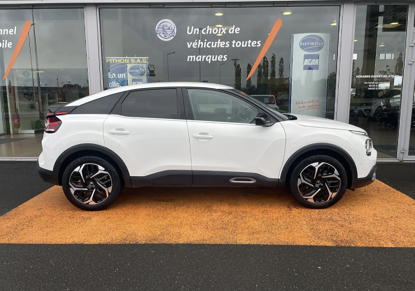 Profil côté gauche d'une Citroën C4 blanche 2022 avec toit noir et jantes alliage distinctives, devant une vitrine commerciale.