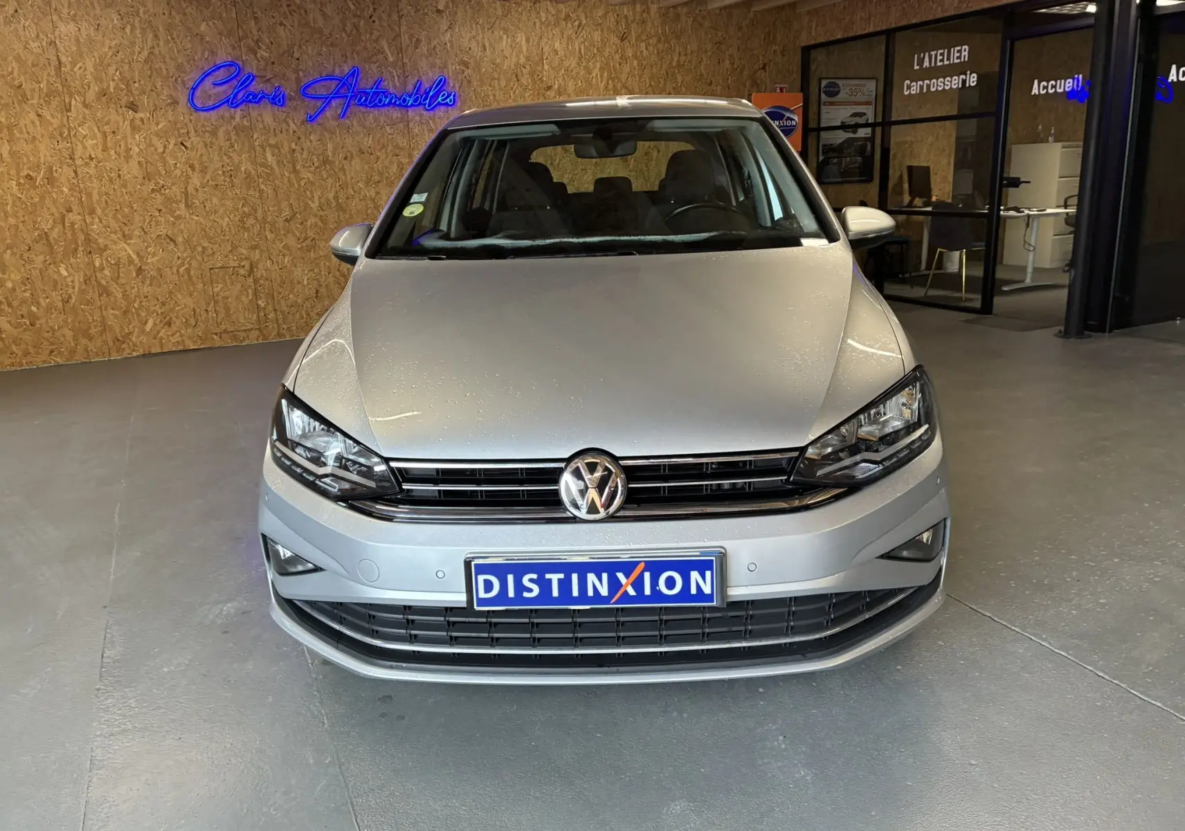 Vue frontale d'une Volkswagen Golf Sportsvan gris clair stationnée en intérieur avec logo Distinxion visible sur la plaque.