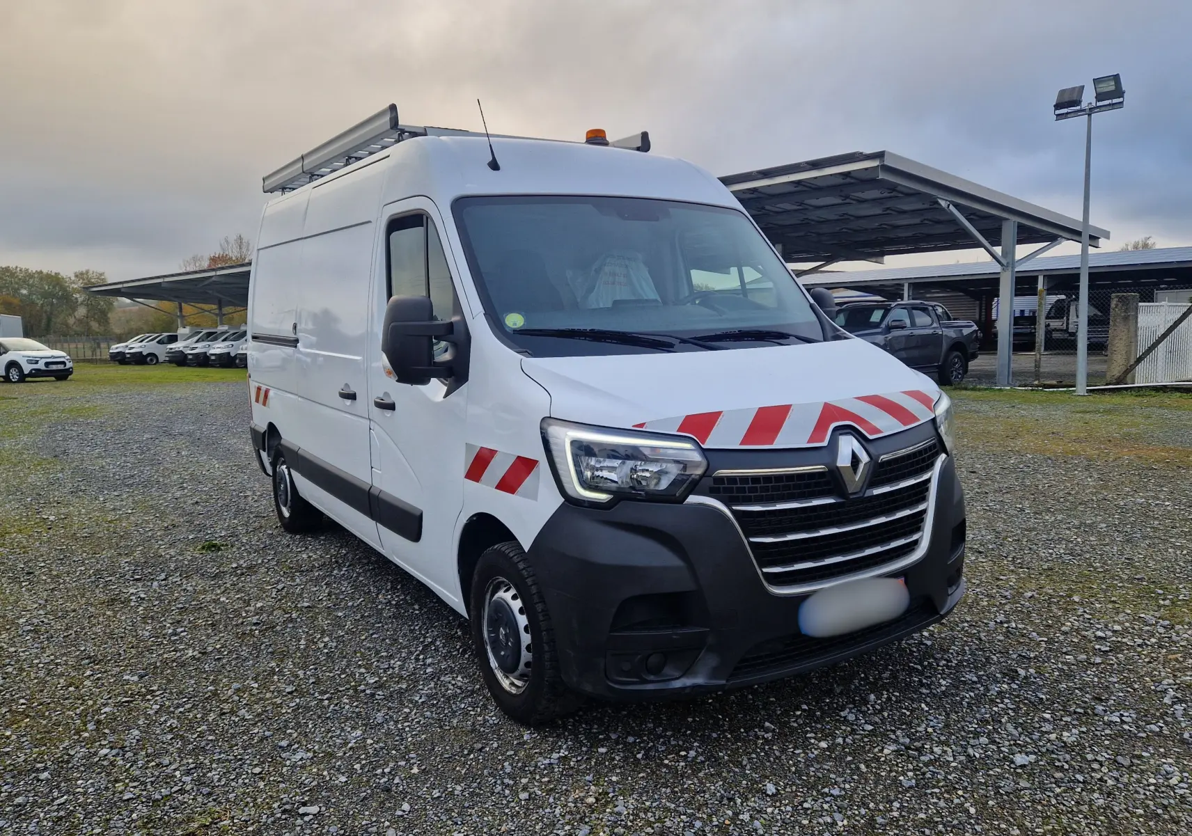 Renault Master blanc en 3/4 avant droit avec bandes rouges réfléchissantes et galerie sur le toit.