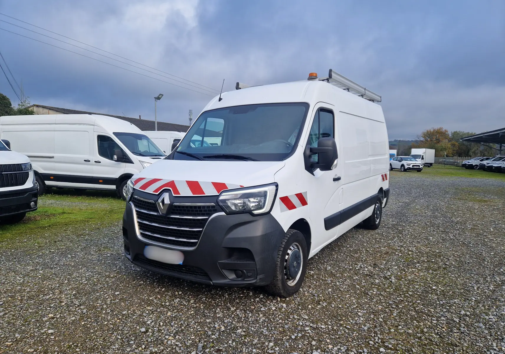 Renault Master blanc en 3/4 avant droit avec bandes rouges réfléchissantes et galerie sur le toit.