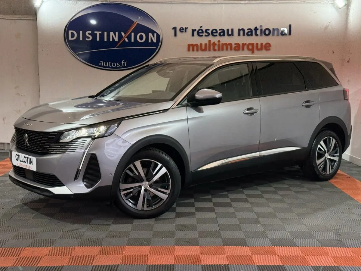 Peugeot 5008 gris clair en vue 3/4 avant droit, avec jantes alliage et phares LED allumés en intérieur showroom.