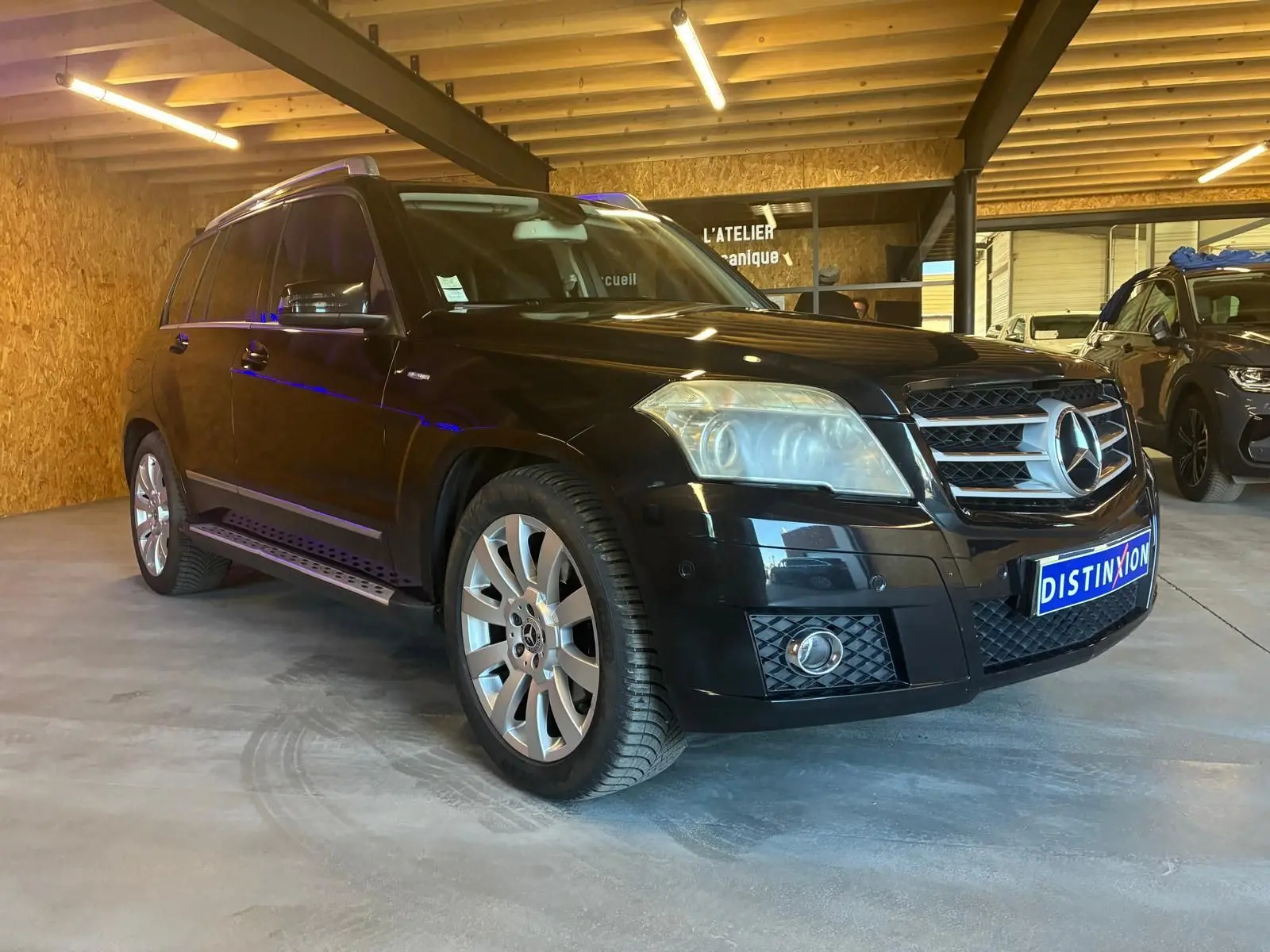 Mercedes GLK noir en 3/4 avant droit dans un garage, avec jantes alliage et calandre chromée visible.