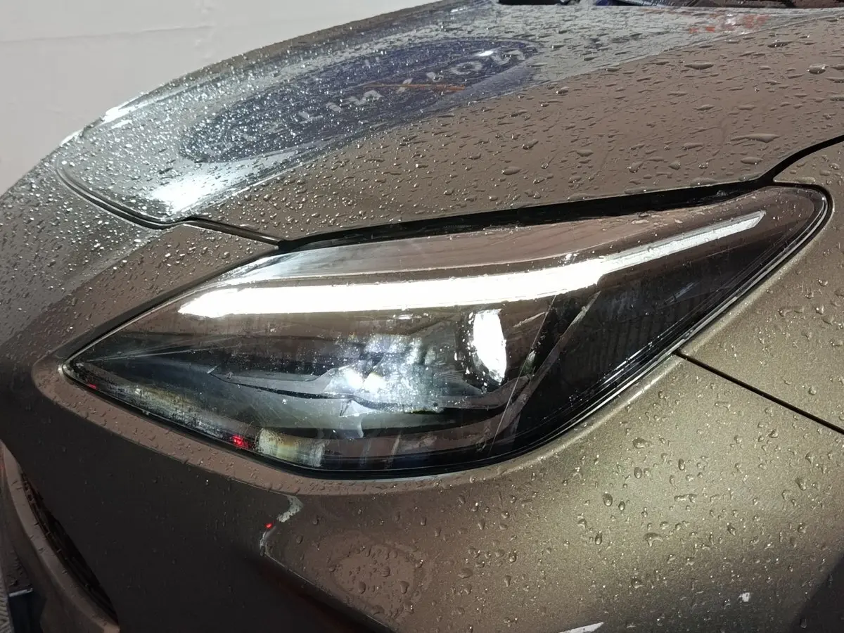 Gros plan sur le phare avant droit allumé d'une Toyota Yaris Cross gris foncé, avec gouttes d'eau sur la carrosserie.