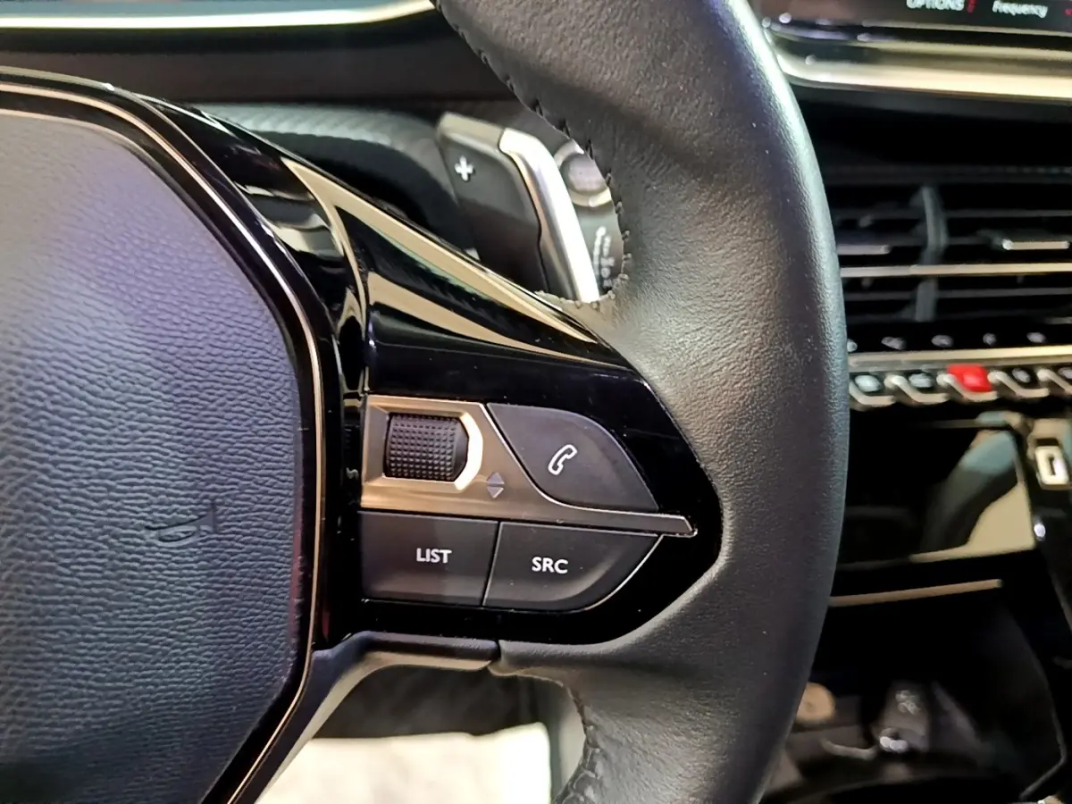 Gros plan sur le volant cuir noir de la Peugeot 2008, avec commandes multifonctions et tableau de bord en arrière-plan.