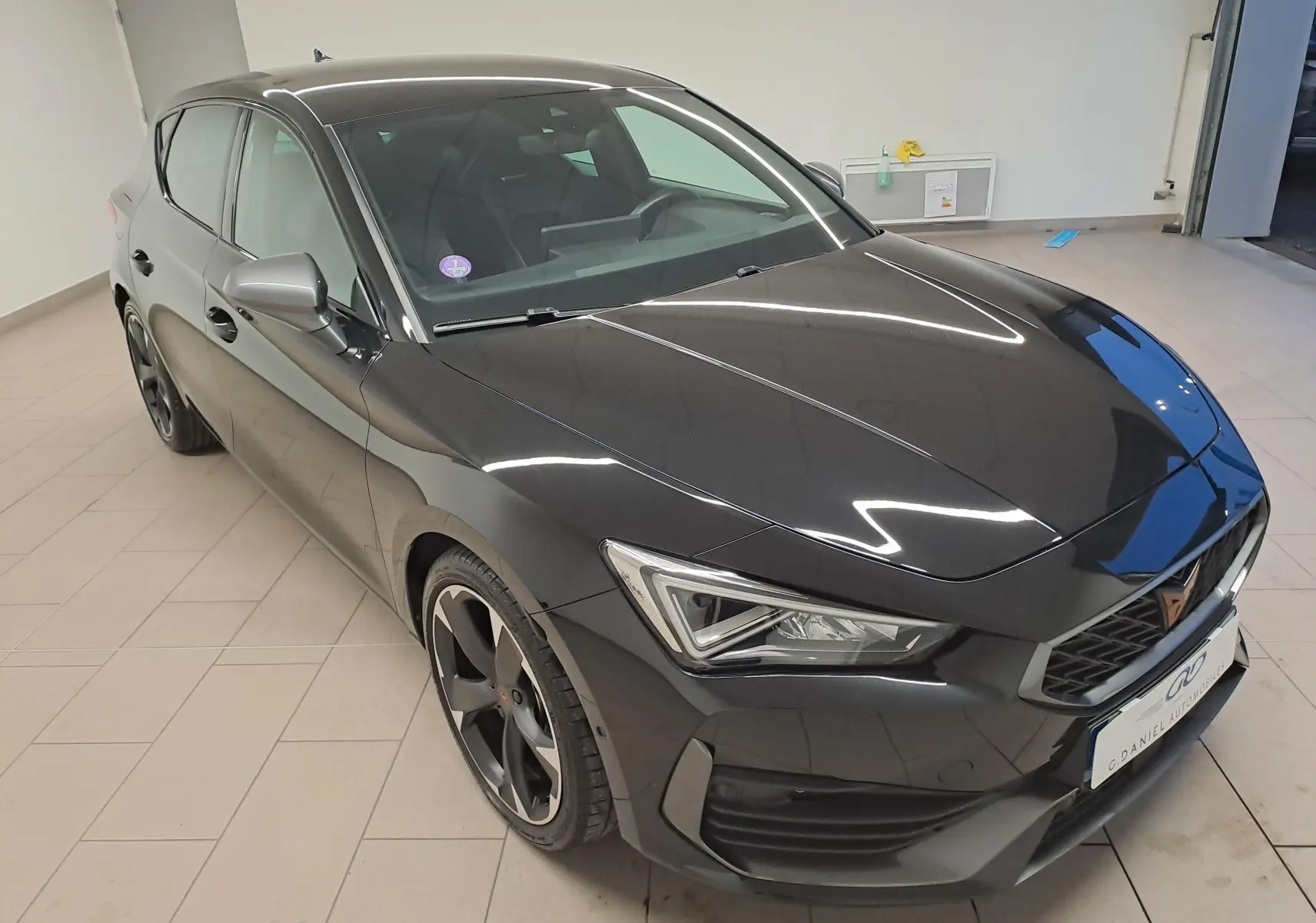 Vue 3/4 avant droit d'une CUPRA Leon 2023 noire métallisée avec jantes noires et calandre distinctive en intérieur showroom.