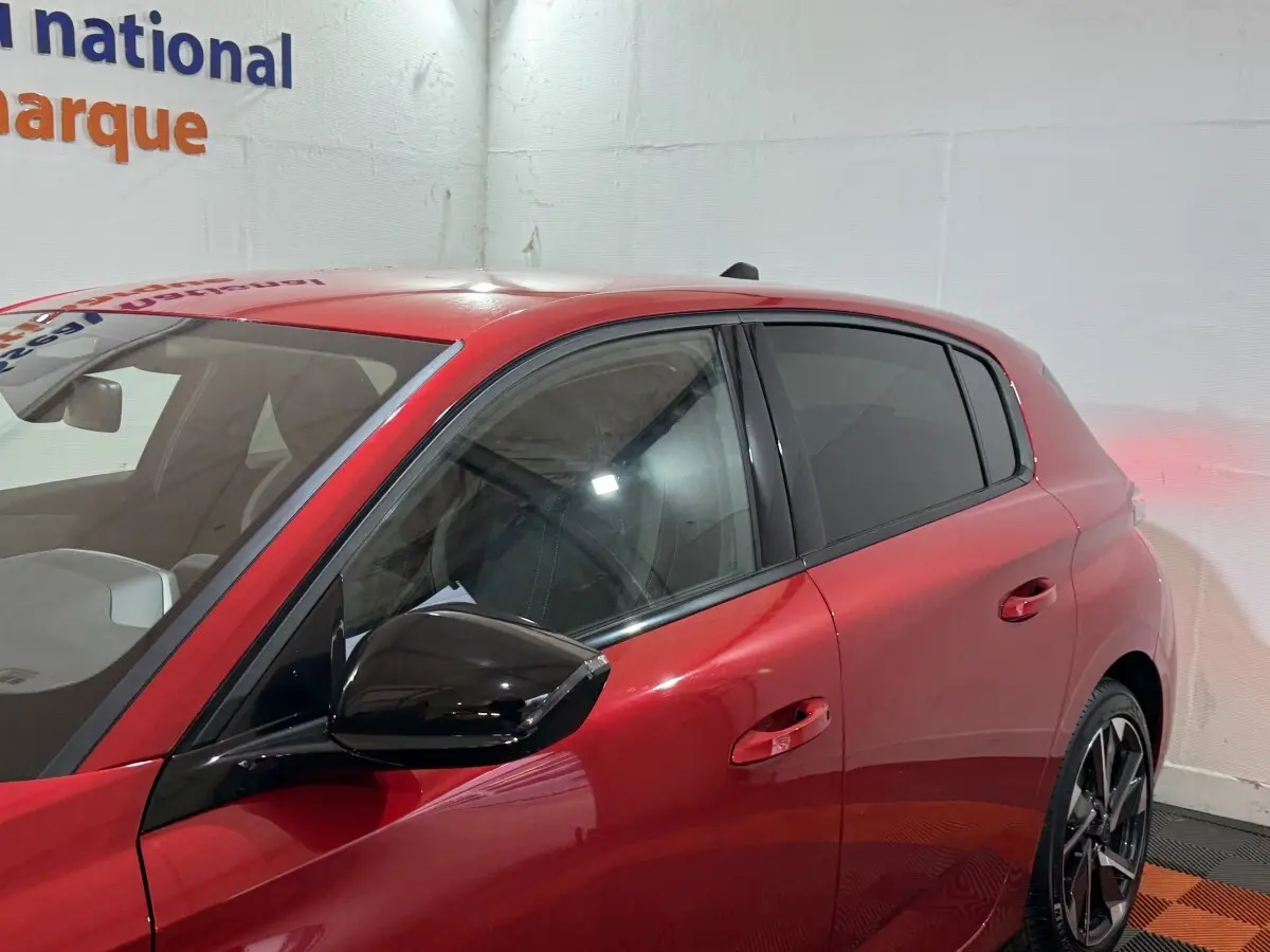 Vue côté gauche avant d'une Peugeot 308 rouge 2025 avec rétroviseur noir brillant et vitres teintées.