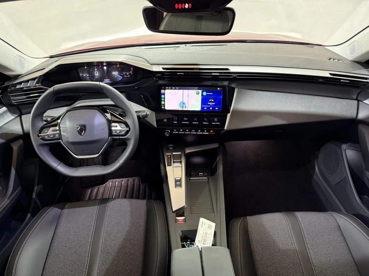 Vue intérieure avant de la Peugeot 308 rouge 2025, volant cuir multifonctions et écran tactile central affichant la navigation.