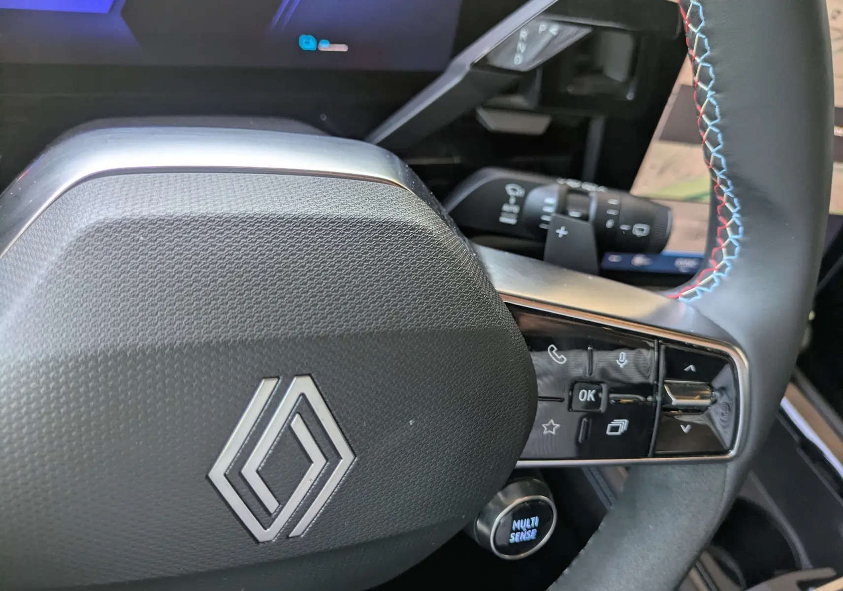 Gros plan sur le volant noir avec logo Renault, commandes intégrées et surpiqûres tricolores visibles à droite.
