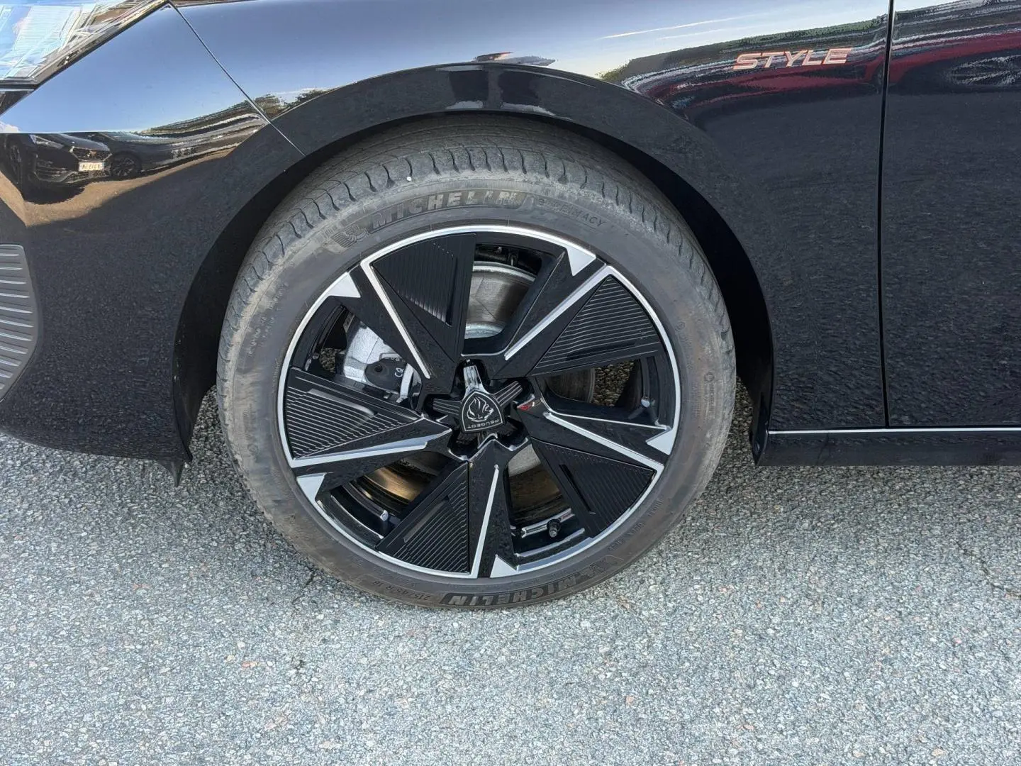 Gros plan sur la roue avant gauche noire avec jante alu design et logo Peugeot, sur carrosserie noire Perla Nera Style.