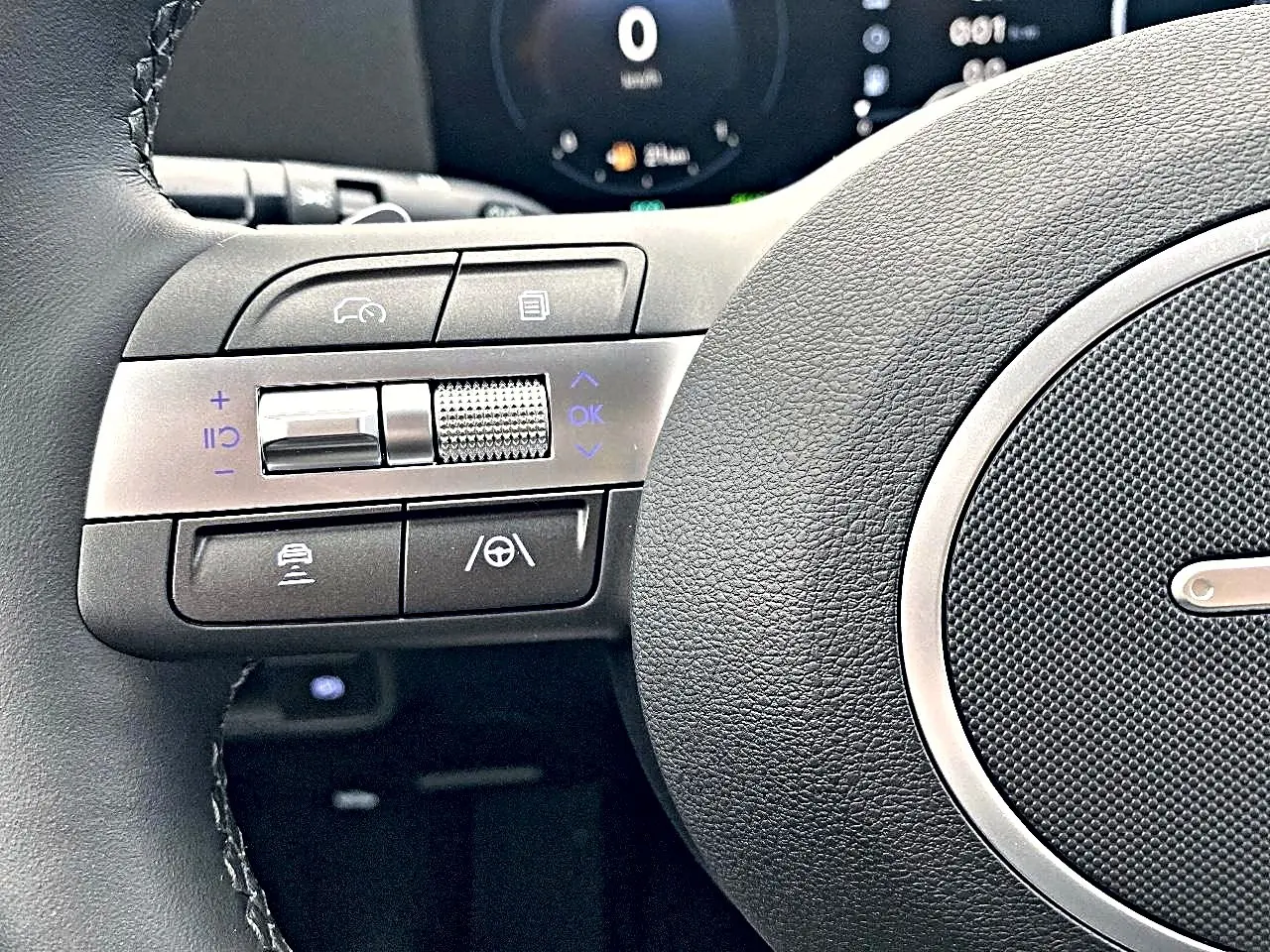 Gros plan sur les commandes à gauche du volant cuir noir du Hyundai Kona gris, avec tableau de bord numérique en arrière-plan.