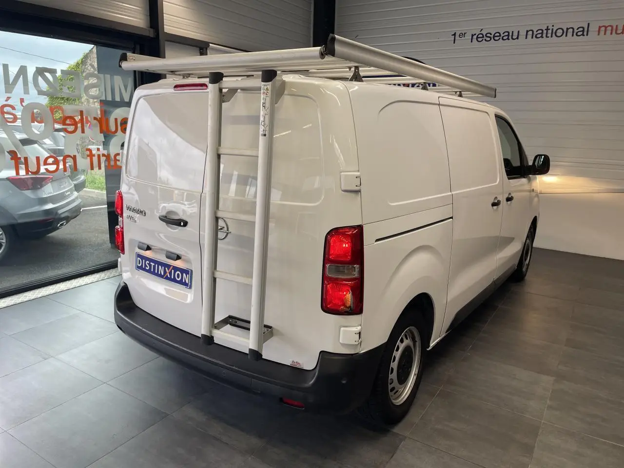 Vue 3/4 arrière droite d'un Opel Vivaro blanc avec échelle et galerie de toit dans un showroom.