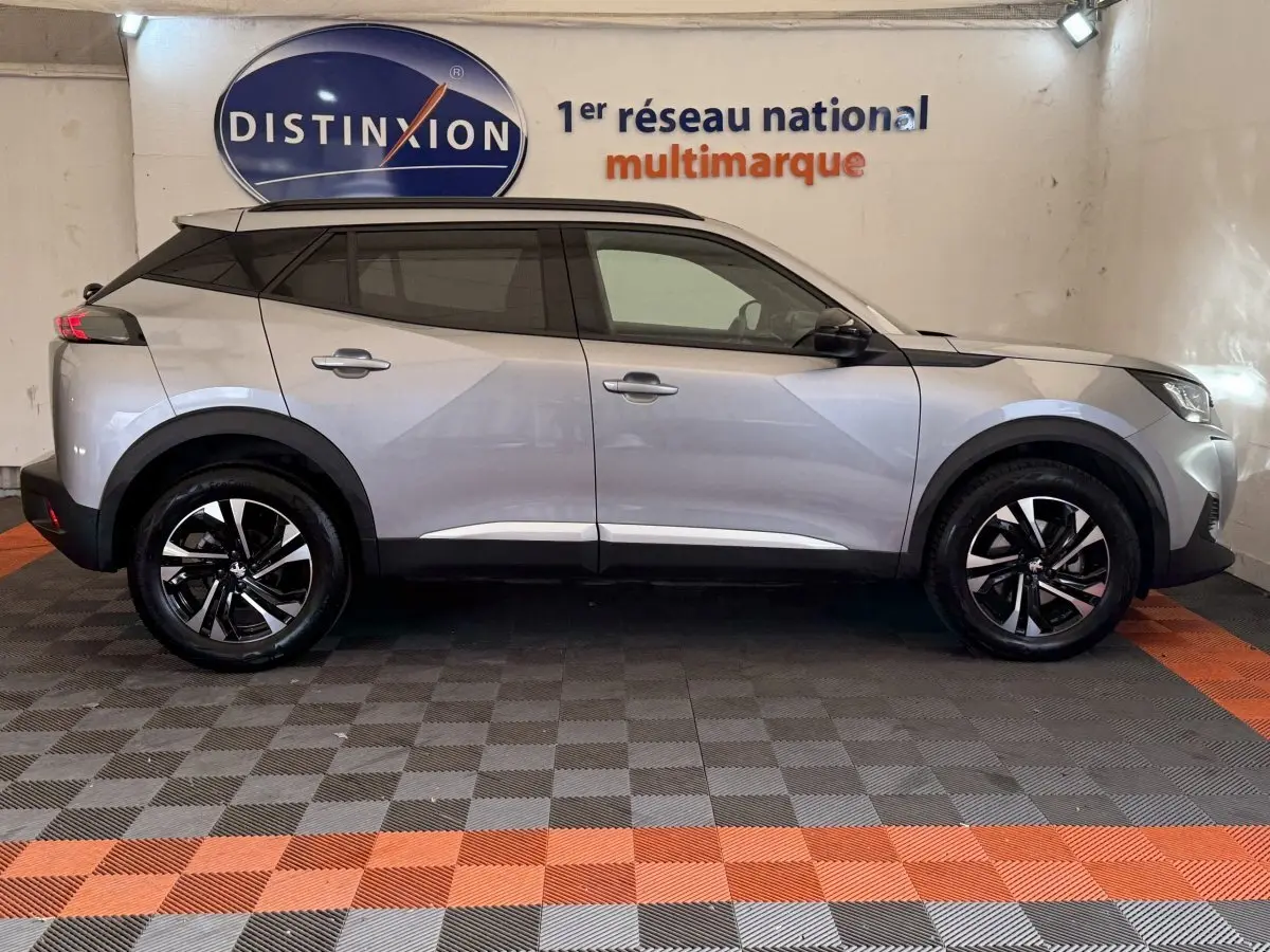 Profil droit d'un Peugeot 2008 gris clair 2023, avec jantes noires et toit noir, stationné en intérieur sur sol damier.