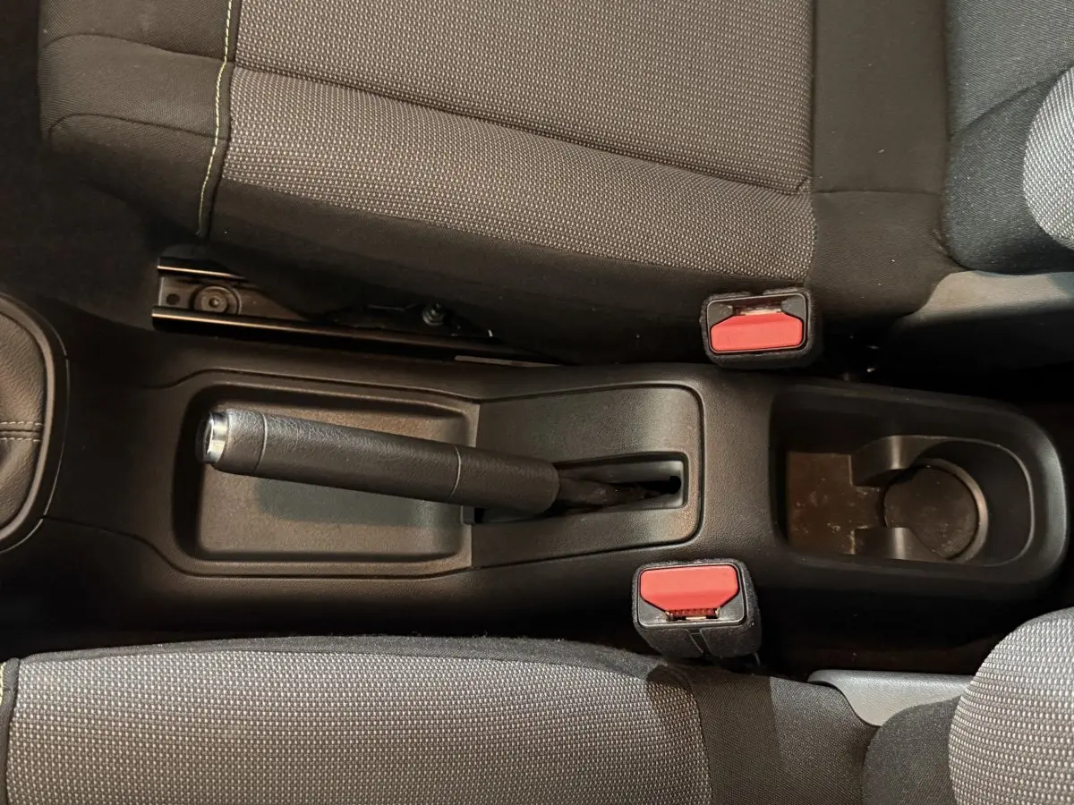 Vue rapprochée de la console centrale avec frein à main manuel et ceintures rouges entre les sièges avant tissu gris.