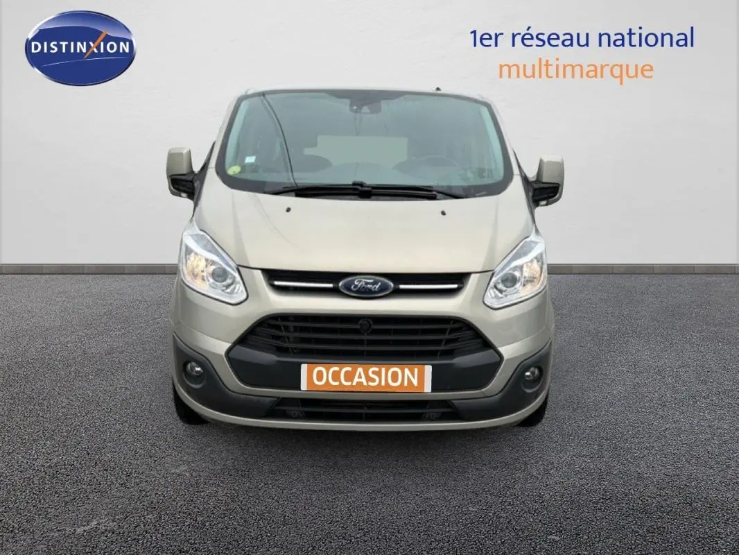 Vue de face d'un Ford Tourneo Custom gris clair avec plaque "OCCASION" et phares allumés sur fond neutre.