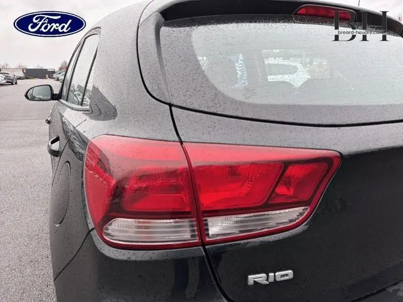 Gros plan sur le feu arrière droit d'une KIA Rio noire Ebène sous la pluie, avec le logo RIO visible sur le hayon.