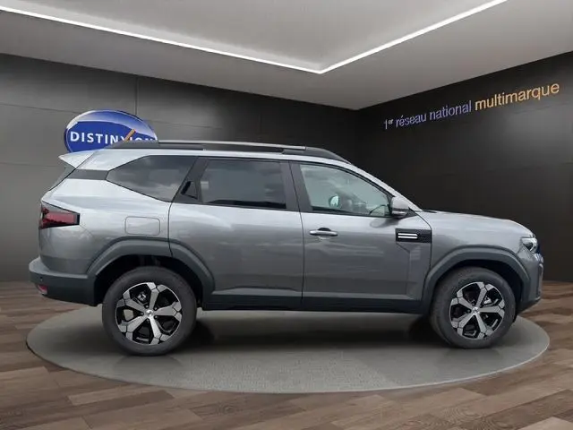 Vue de profil côté droit d'un Dacia Bigster gris foncé avec jantes alliage et barres de toit noires en intérieur showroom.