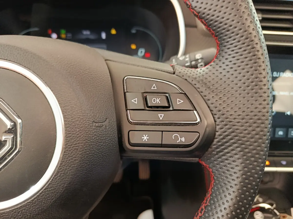 Gros plan sur le volant noir perforé avec surpiqûres rouges du MG ZS Luxury 2024, boutons de commande multifonctions visibles.