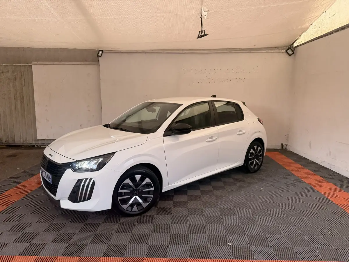 Peugeot 208 blanche vue en 3/4 avant droit, avec jantes noires et phares LED allumés, stationnée en intérieur.