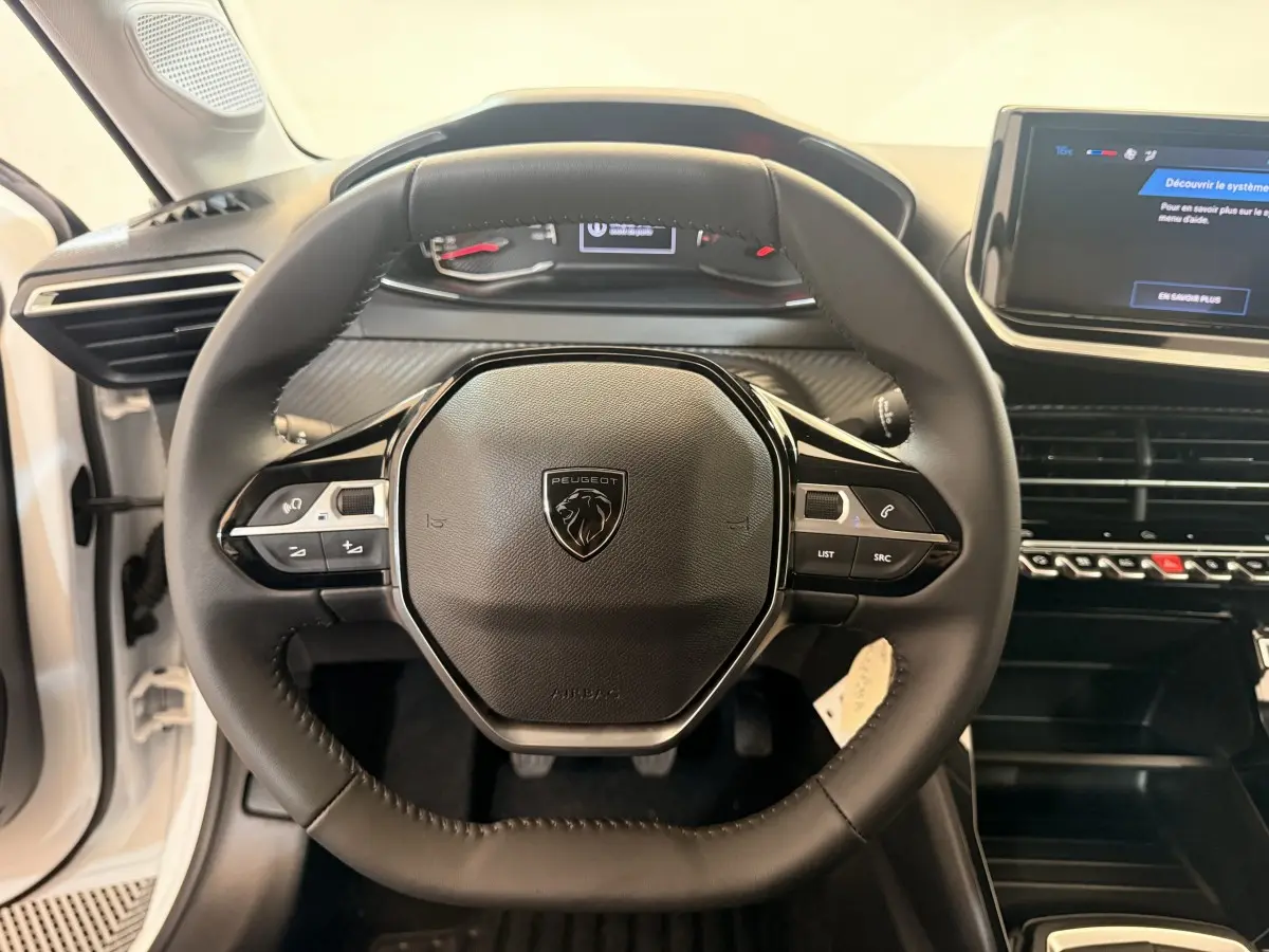 Vue rapprochée du volant cuir noir de la Peugeot 208 blanche, avec tableau de bord digital et écran tactile central.