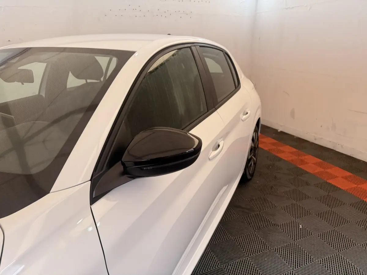 Vue côté gauche arrière d'une Peugeot 208 blanche avec rétroviseur noir et vitres teintées dans un intérieur fermé.