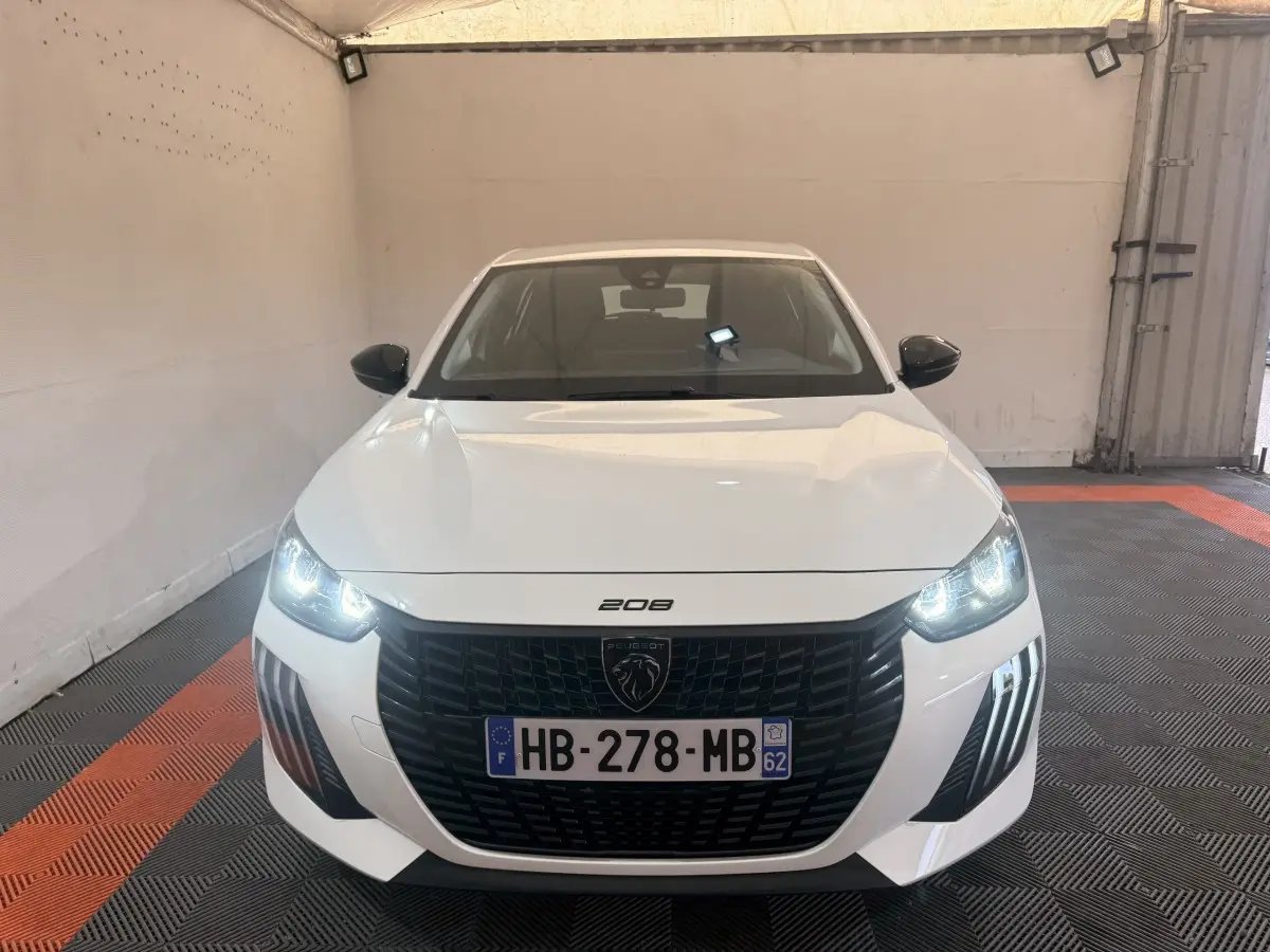 Vue de face d'une Peugeot 208 blanche avec phares allumés et calandre noire dans un garage.