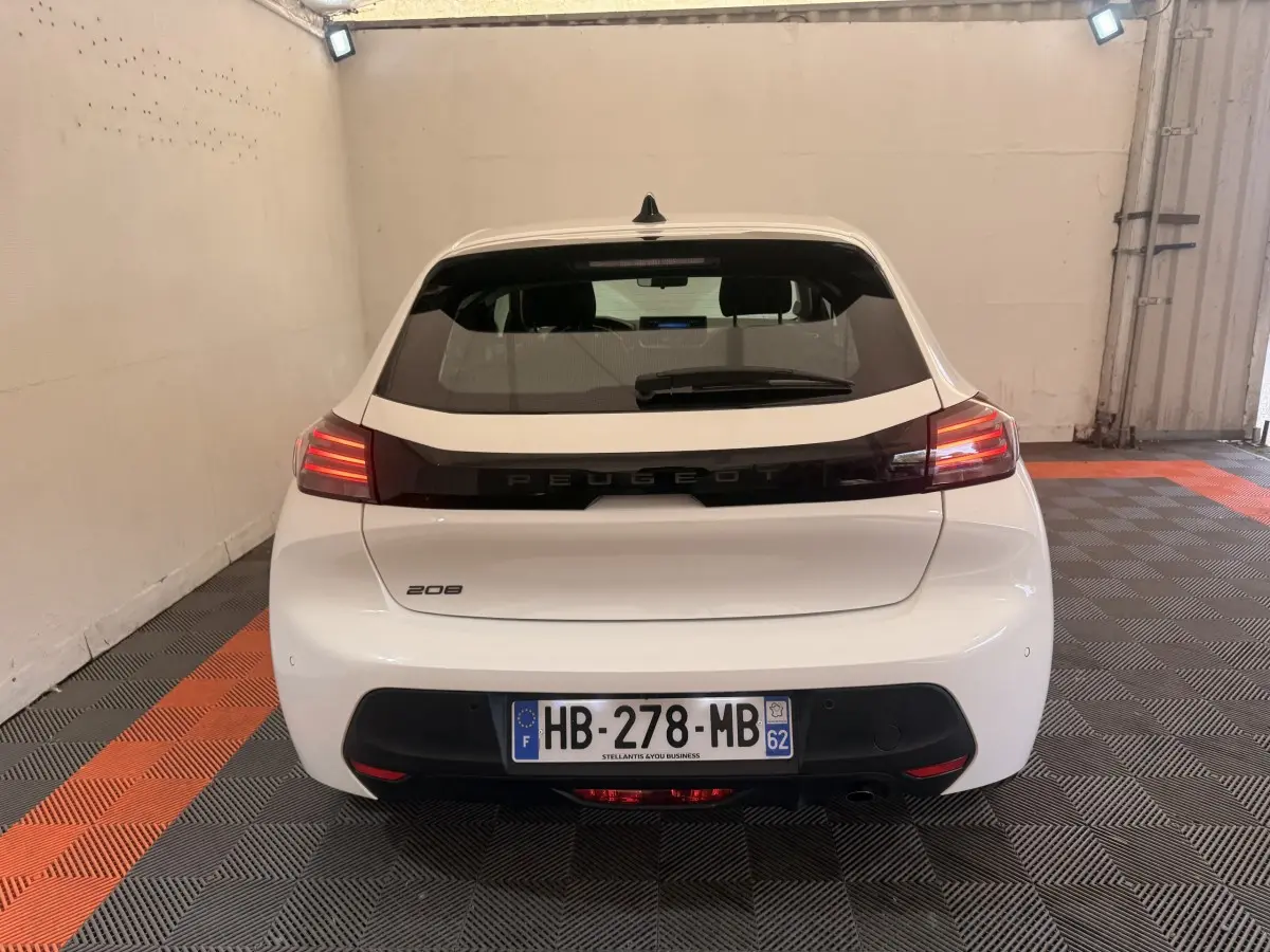 Vue arrière d'une Peugeot 208 blanche 2025 dans un garage, avec feux arrière allumés et plaque d'immatriculation visible.