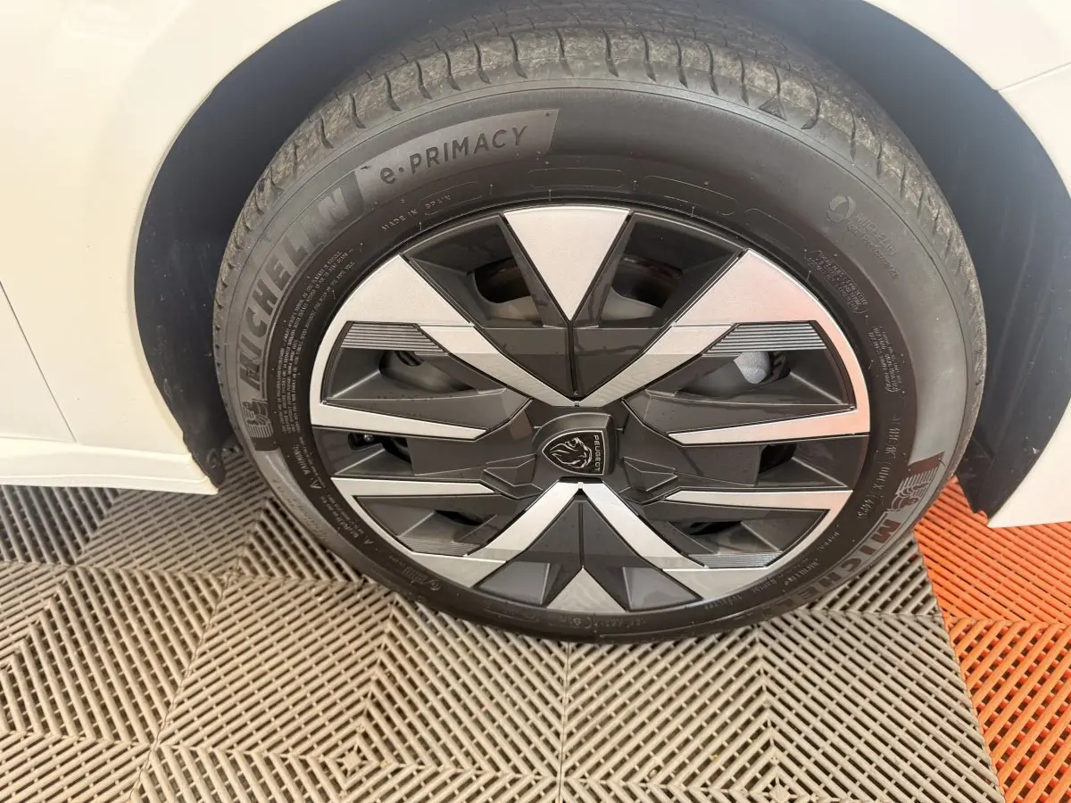 Gros plan sur la roue avant gauche d'une Peugeot 208 blanche, avec jante au design géométrique noir et argent.