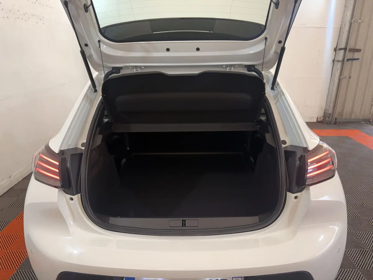 Vue arrière ouverte du coffre d'une Peugeot 208 blanche 2025 avec feux arrière LED allumés dans un garage.