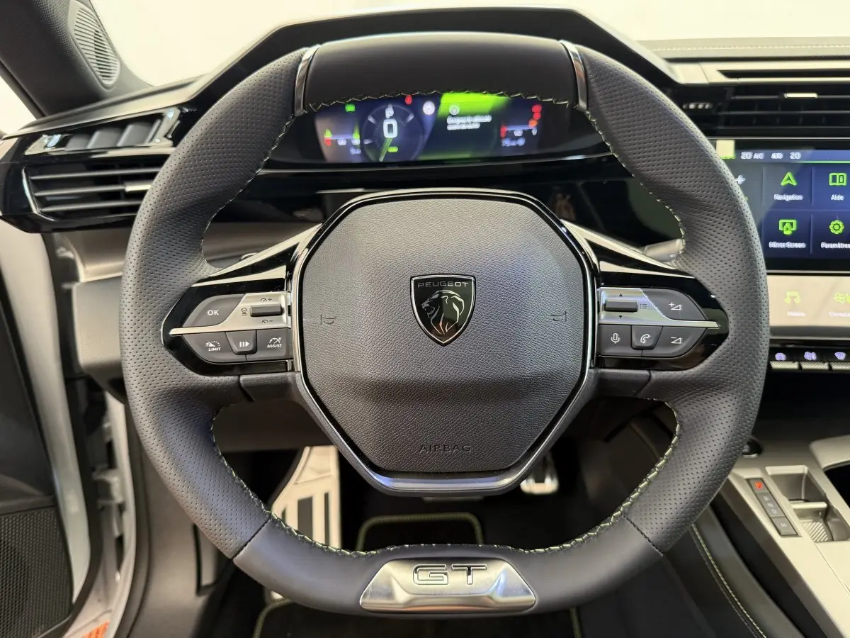 Vue rapprochée du volant cuir noir avec logo Peugeot et badge GT, tableau de bord digital et écran tactile visibles.